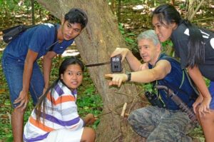 Four people attach a wildlife camera to a tree. They are all crouching at the trunk of a tree. Dr Kyes is pointing to something out of field.